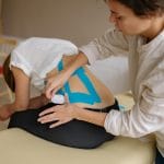 Jak trenować stabilizację po epizodzie rwy kulszowej: zasady i bezpieczne ćwiczenia Fizjoterapeuta nakleja niebieskie taśmy kinesiotaping na plecy pacjenta