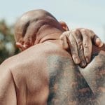 Łysy mężczyzna z tatuażem trzyma się za bolące plecy na słońcu